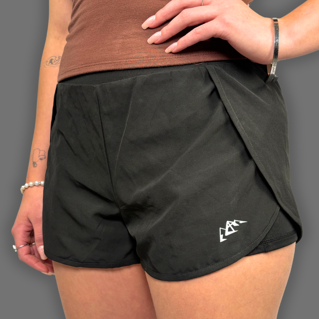 Black athletic shorts with a side flap detail and a small white logo, shown on a model against a solid gray background.
