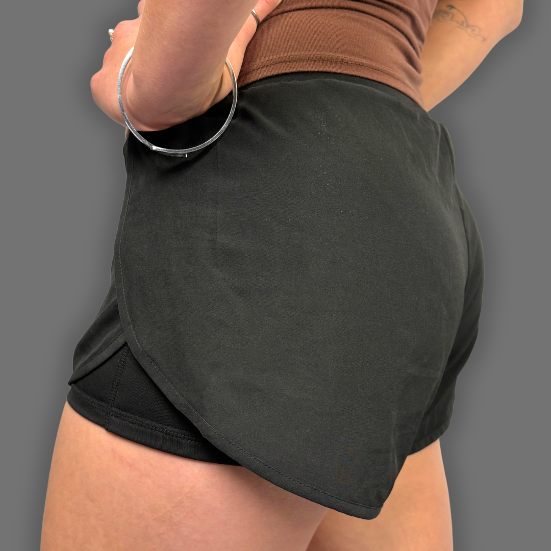 A close-up of a person wearing a dark charcoal gray wrap-style athletic skirt over black shorts, paired with a brown top, shown from the back against a solid gray background.