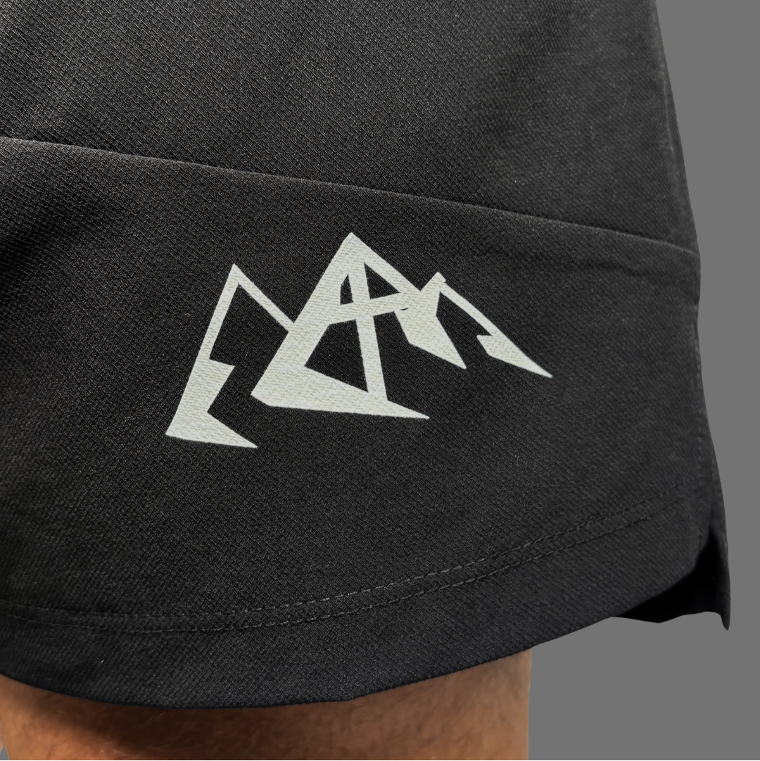 Close-up of a black athletic shorts leg featuring a white geometric mountain logo on the upper thigh, with a person's skin visible at the bottom edge.