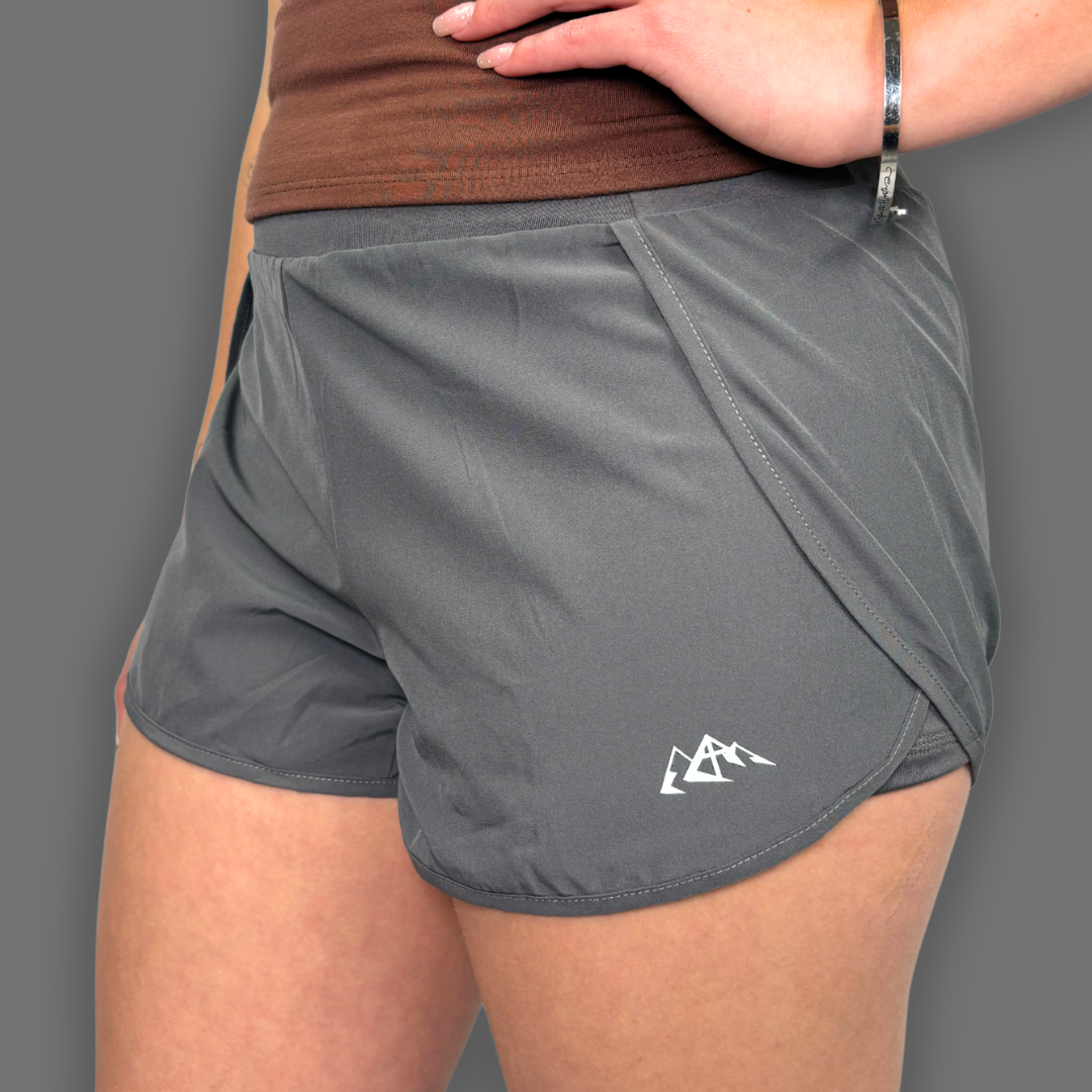 A close-up of a person wearing gray athletic shorts with a white mountain logo near the hem, paired with a brown top against a solid gray background.