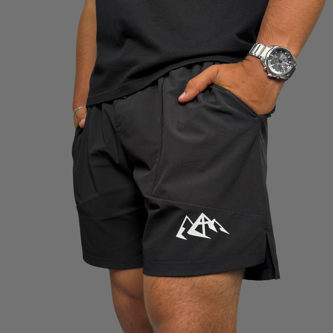 A close-up of a person wearing black athletic shorts with a white mountain logo on the lower left leg, paired with a black t-shirt and a silver watch, against a solid gray background.