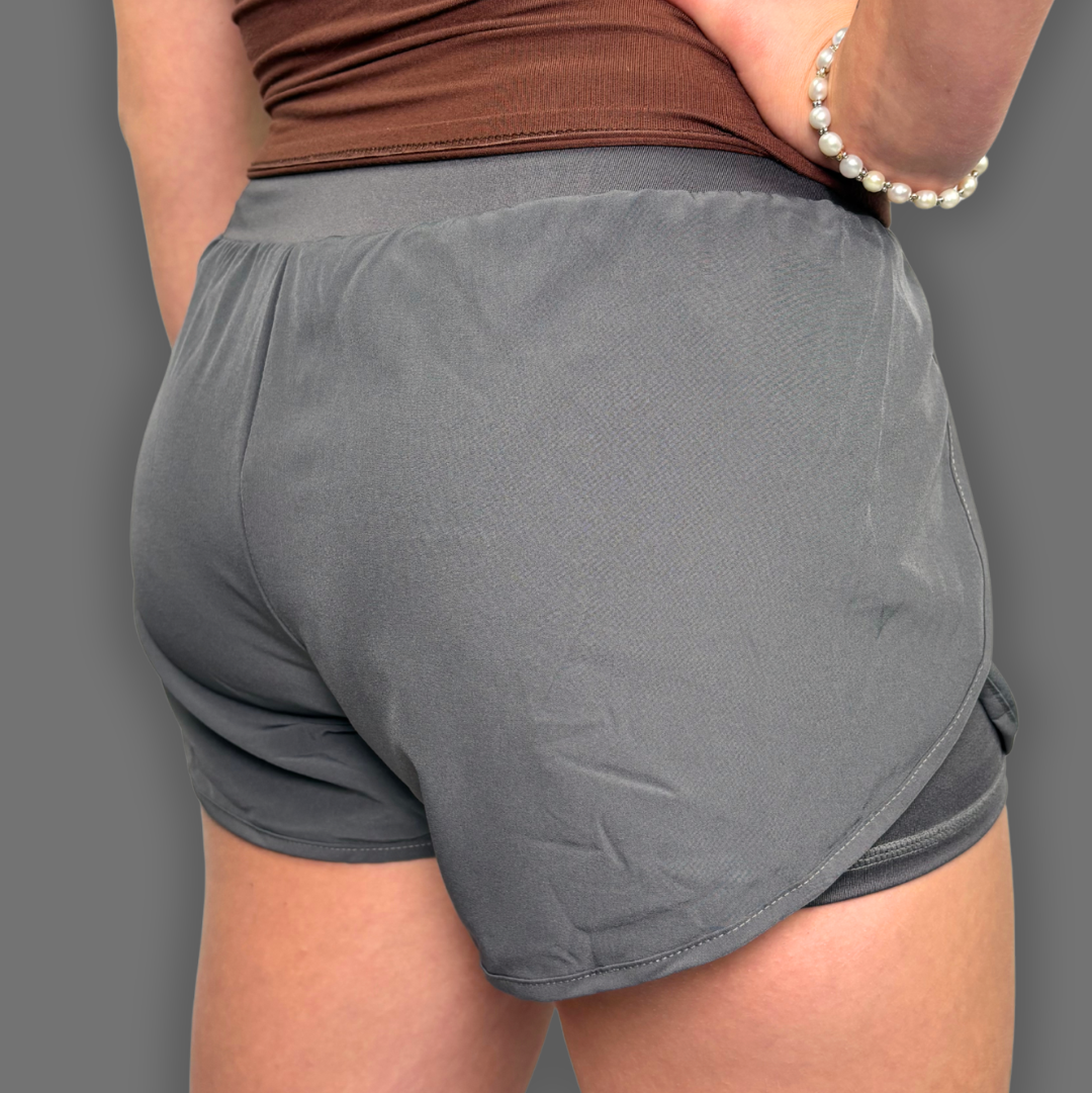 A close-up back view of a person wearing gray athletic shorts and a brown top, with one hand on the hip and a pearl bracelet visible against a plain gray background.
