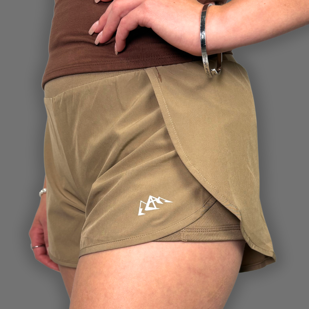 A close-up of a person wearing tan athletic shorts with a white mountain logo on the left leg, paired with a brown top against a solid gray background.