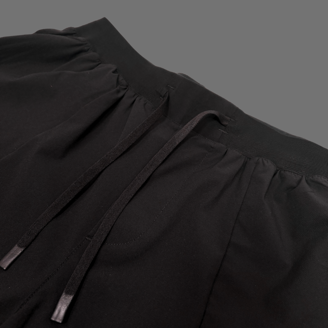 Close-up of a black garment with gathered elastic waistband and dark drawstrings against a solid dark gray background.