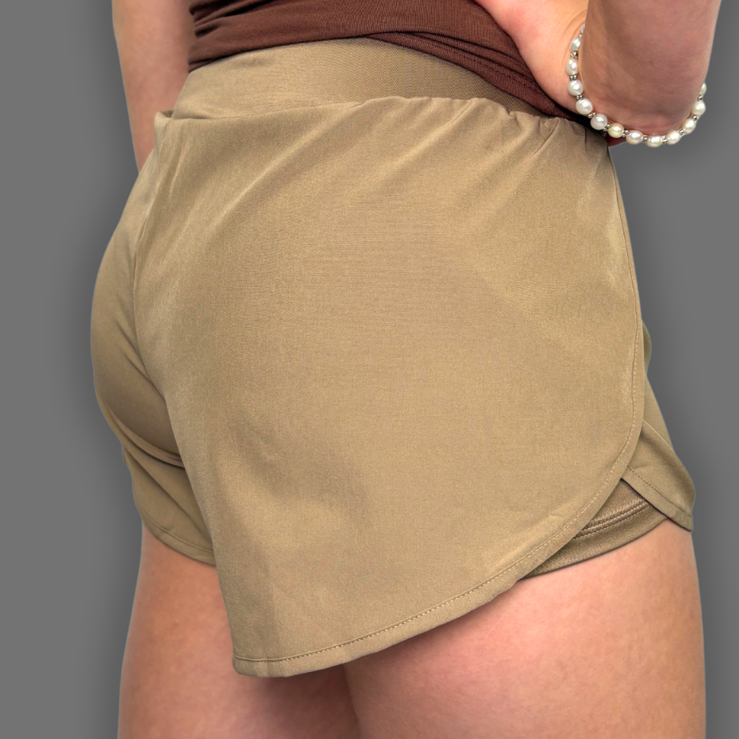 A close-up back view of a person wearing tan athletic shorts and a brown top, with one hand pulling up the shorts. The background is a solid gray studio backdrop.