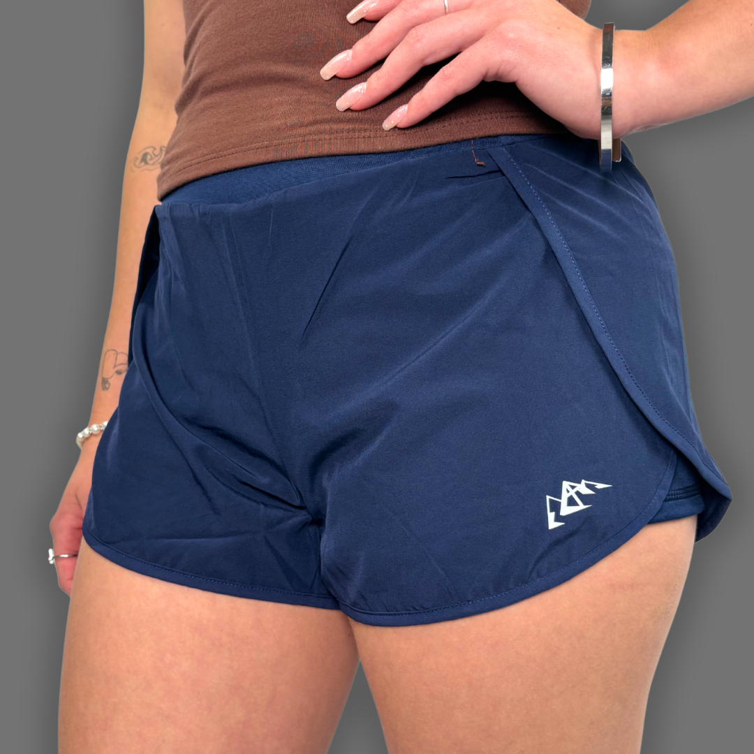 A close-up of a model wearing navy blue athletic shorts with a white logo on the lower left leg, paired with a brown tank top against a solid gray background.