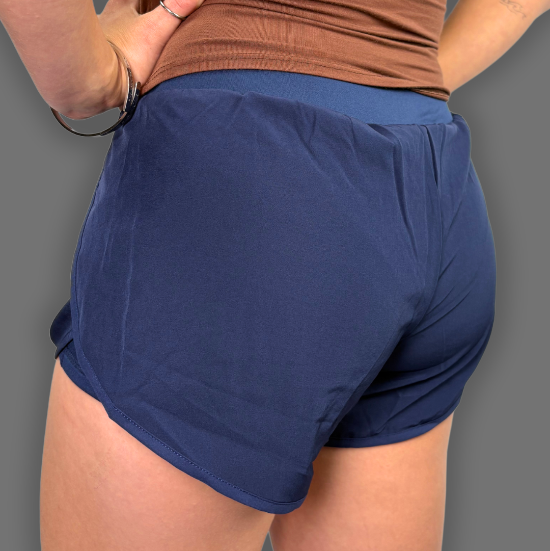 A close-up back view of a person wearing navy blue athletic shorts and a brown ribbed tank top against a solid gray background.