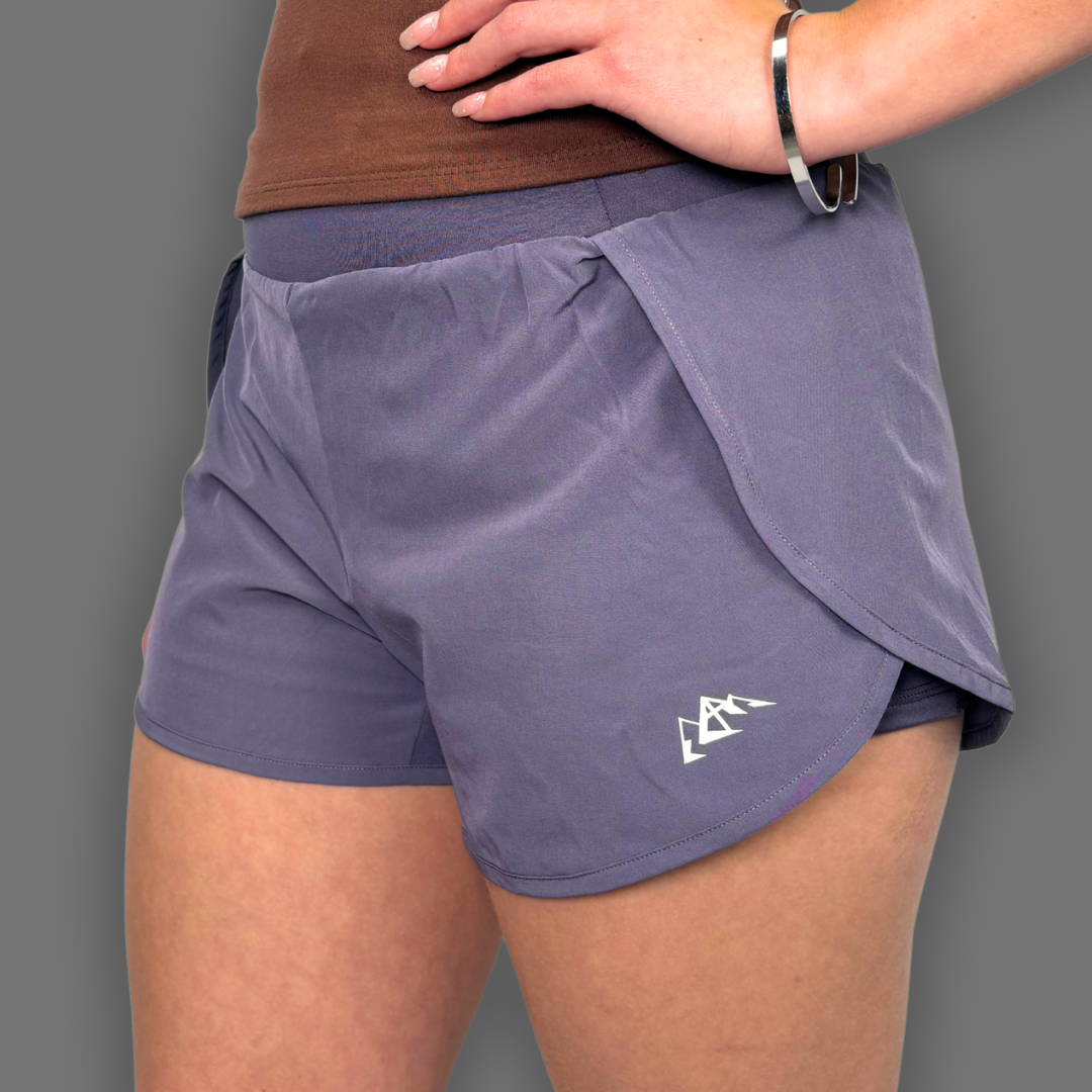 A close-up of a person wearing slate purple athletic shorts with a white logo on the lower left leg, paired with a brown top against a solid gray background.