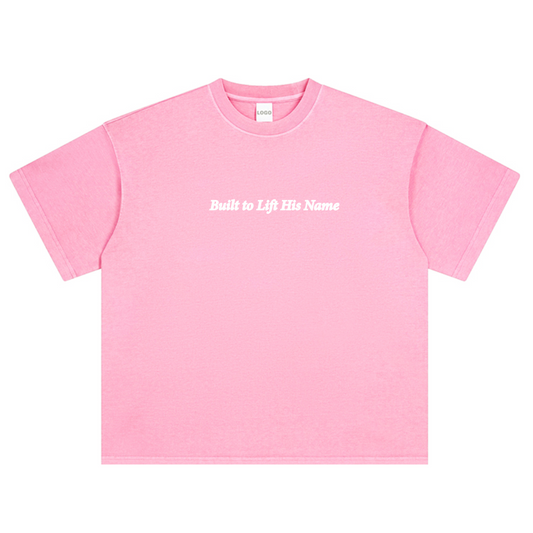 Pink t-shirt with text 'Built to Lift His Name' on a white background