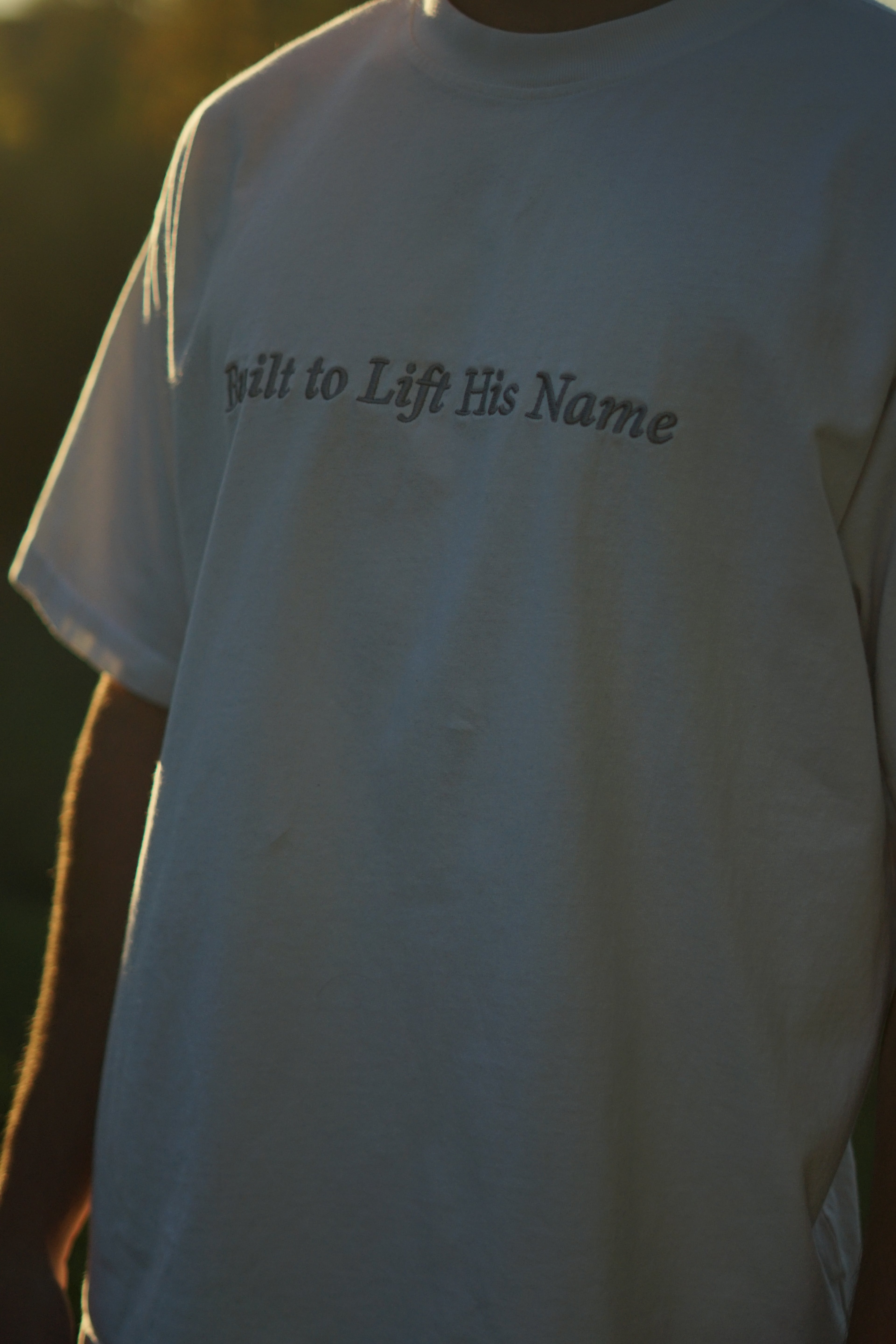 White shirt with text 'Built to Lift His Name' worn by a person, blurred background for Levage