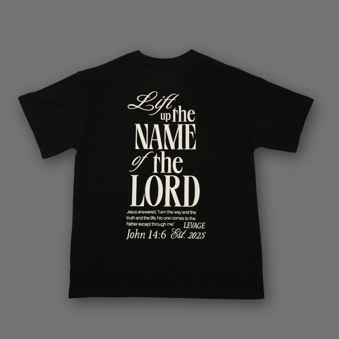 A black short-sleeve t-shirt with a white typographic graphic on the front reading 'Lift up the NAME of the LORD' and smaller scripture text below, shown on a dark gray background.