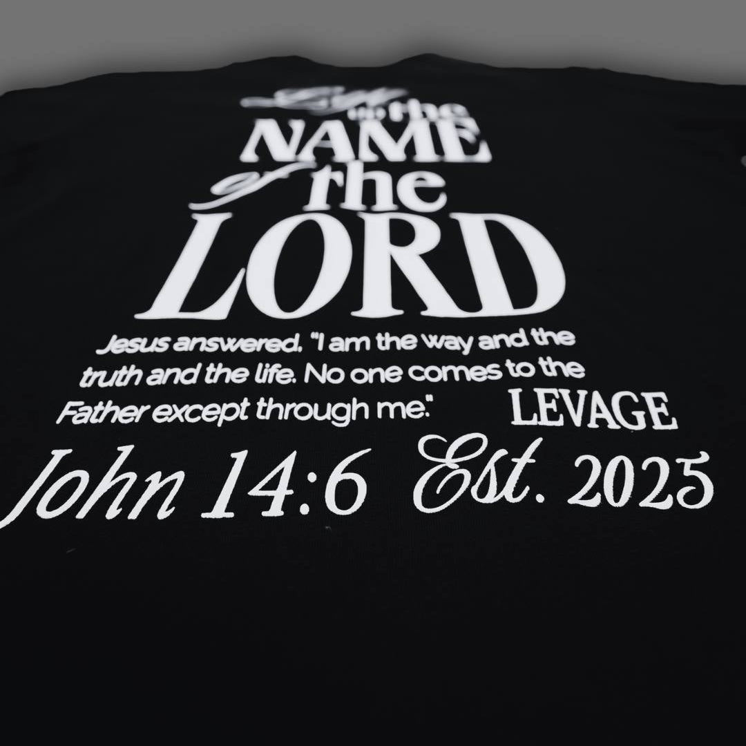 A black t-shirt featuring a white printed graphic with religious text, including 'The NAME of the LORD' and a Bible verse from John 14:6 with 'LEVAGE Est. 2025' written at the bottom.