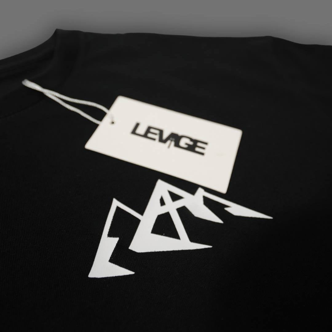 A close-up of a black t-shirt featuring a white geometric mountain graphic and a white paper tag with the brand name 'LEVAGE' printed on it.