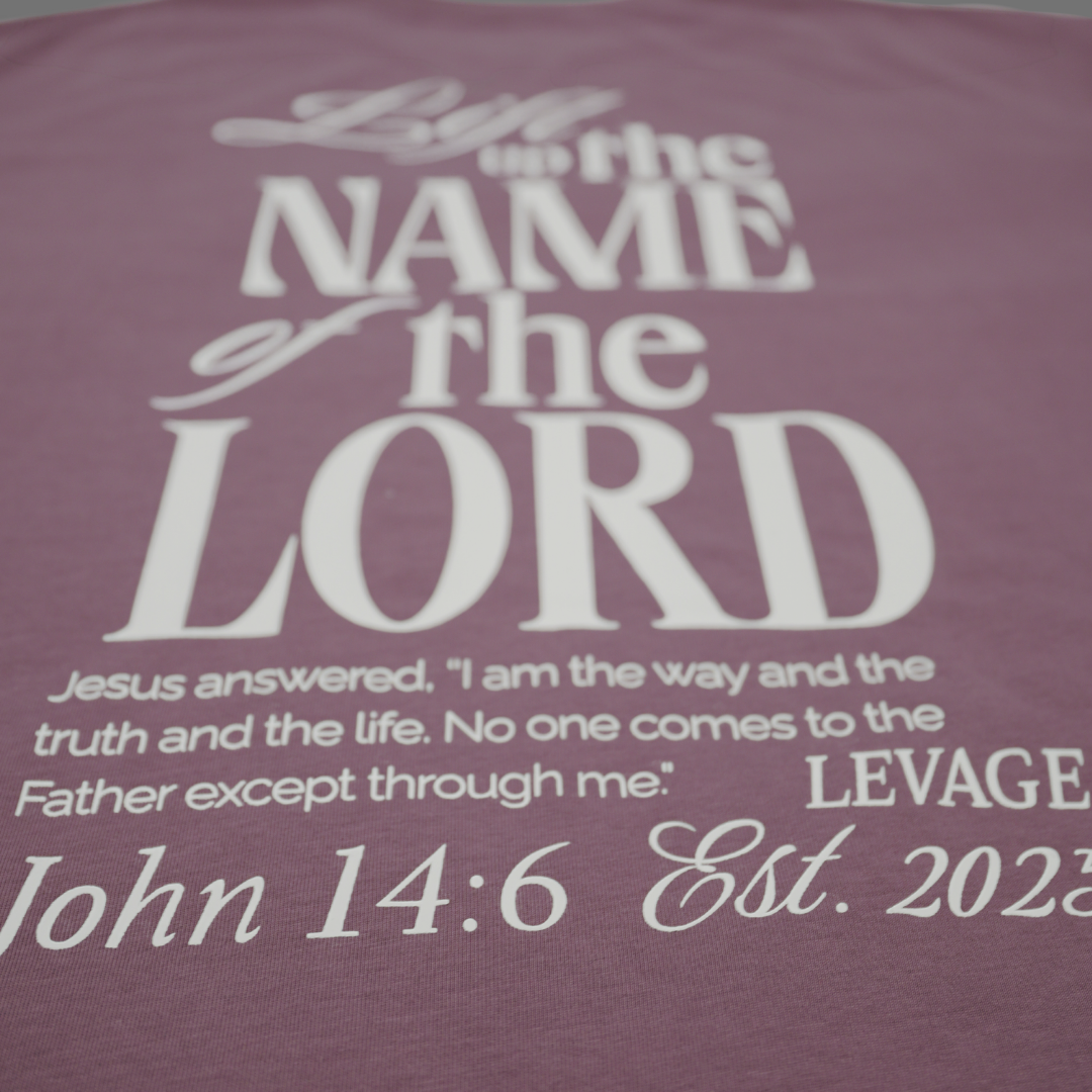 An english lavender t-shirt shown from the back, featuring a tonal white graphic print that reads 'Lift up the name of the Lord' with scripture text below.