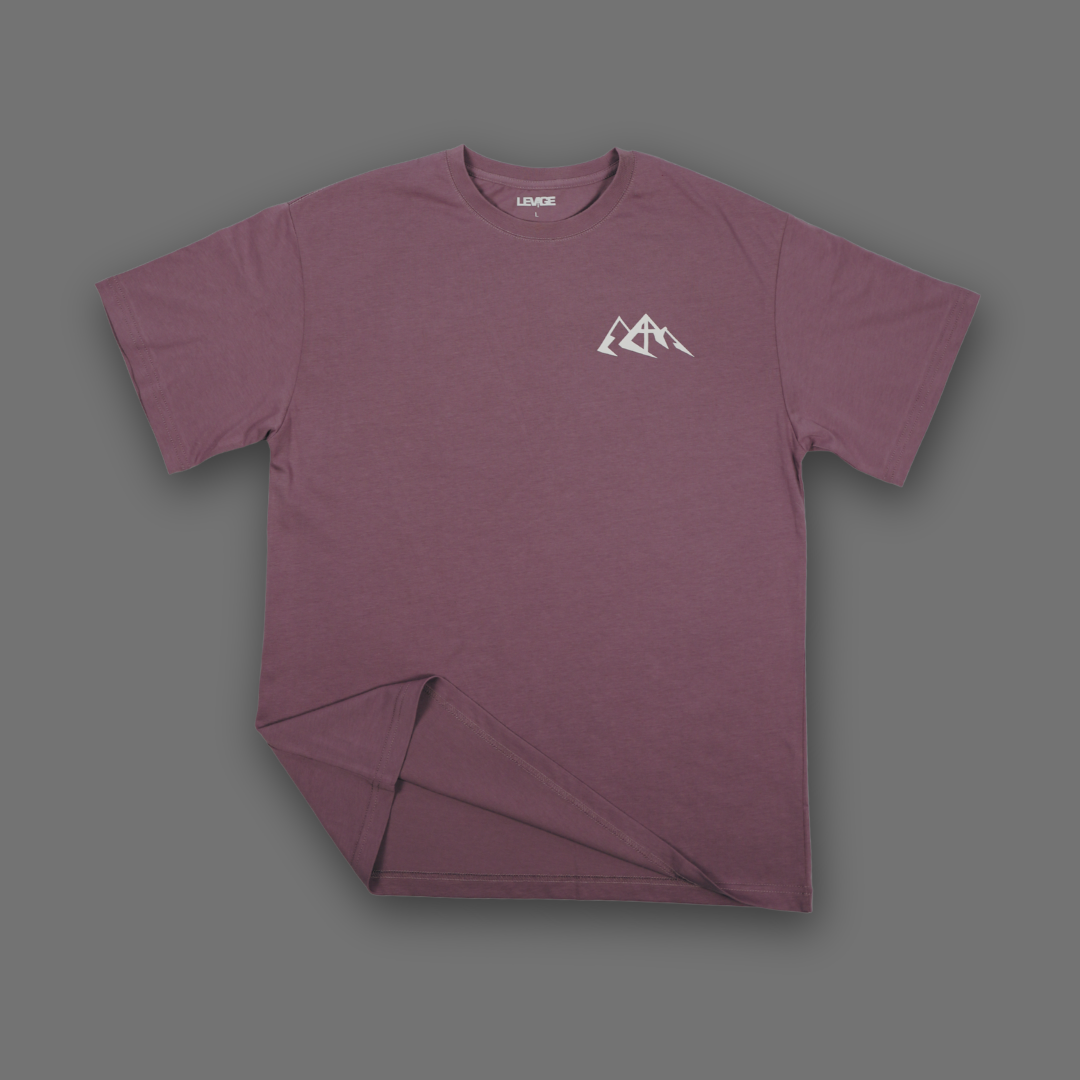 A short-sleeve crewneck t-shirt in an english lavender color, featuring a small white mountain graphic printed on the left chest. The shirt is displayed flat against a solid dark gray background.