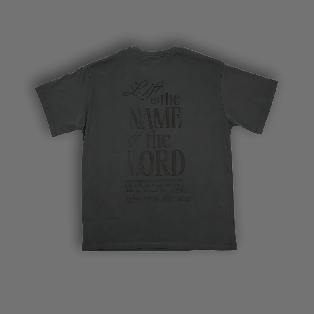 A dark gray t-shirt shown from the back, featuring a tonal black graphic print that reads 'Lift up the name of the Lord' with scripture text below, set against a solid dark gray background.