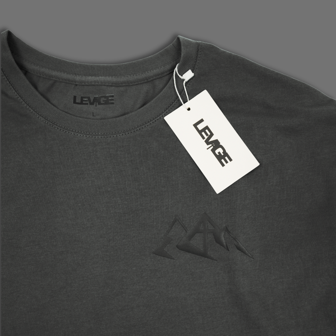 A white short-sleeve t-shirt shown from the front, featuring a black typographic print with the words 'Lift up the NAME of the LORD' and a Bible verse from John 14:6 . The t-shirt is displayed flat against a solid dark gray background with a hangtag.