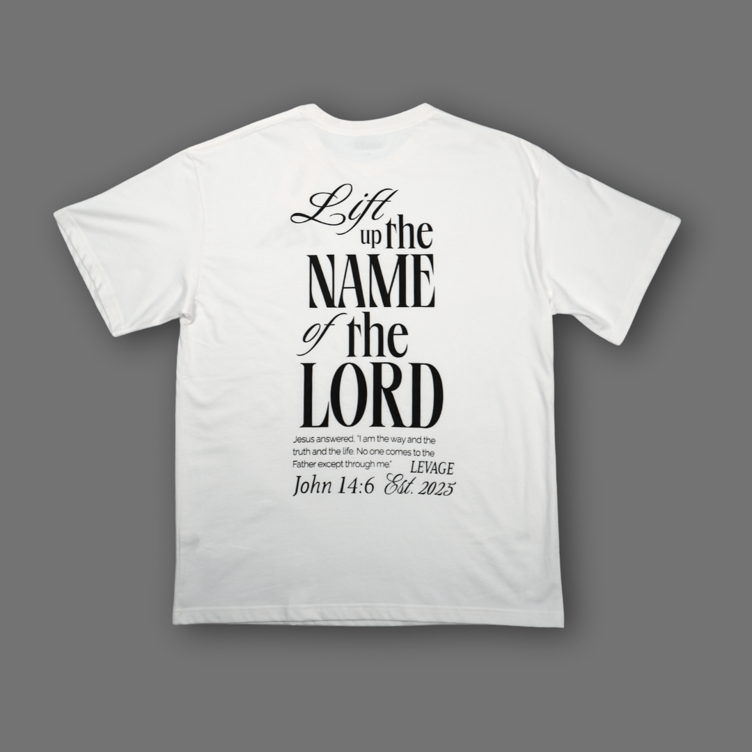 A white short-sleeve t-shirt shown from the back, featuring a black typographic print with the text 'Lift up the NAME of the LORD' and a Bible verse from John 14:6 Levage Est. 2025. The t-shirt is displayed flat against a solid dark gray background.